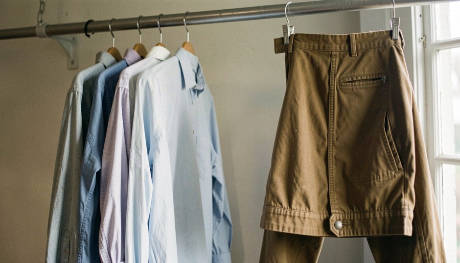 A short row of plain work shirts and one waxed jacket hangs on a peg rail above folded canvas trousers.