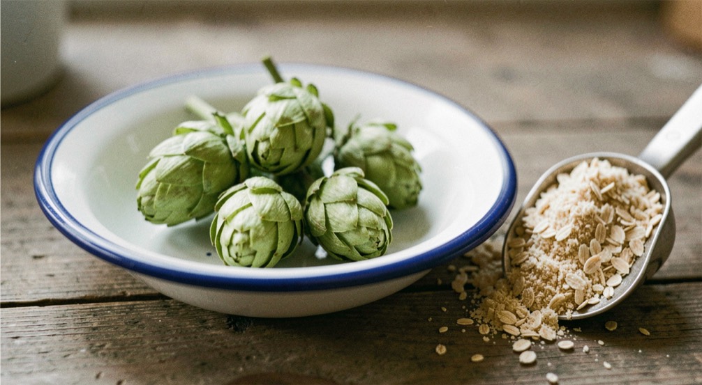 Green hop cones rest in a small enamel dish beside a scoop of crushed malt.